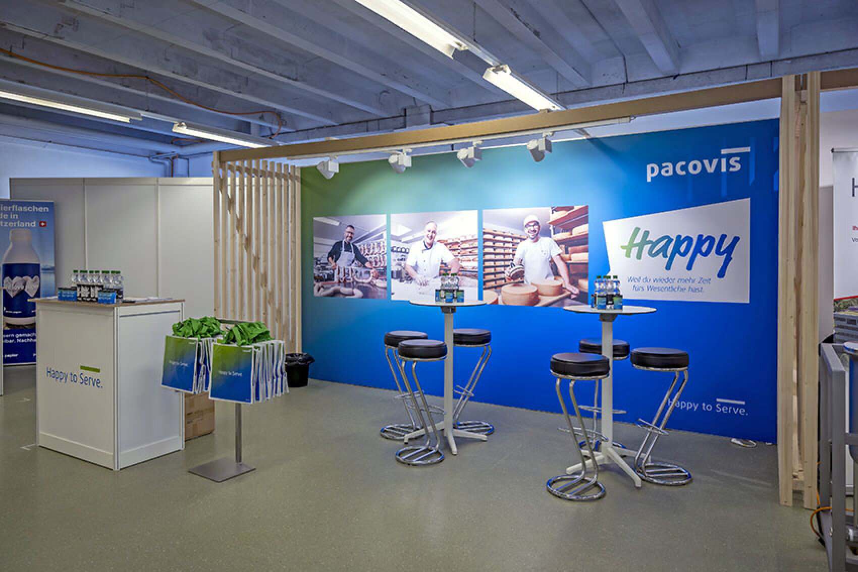 Ein Messestand von Pacovis mit einer Empfangsstheke im Marken-Design, grünen Einkaufstaschen und zwei Stehtischen mit Hockern vor einer blauen Wand, auf der Fotos von der Käseherstellung und das Wort Happy zu sehen sind.