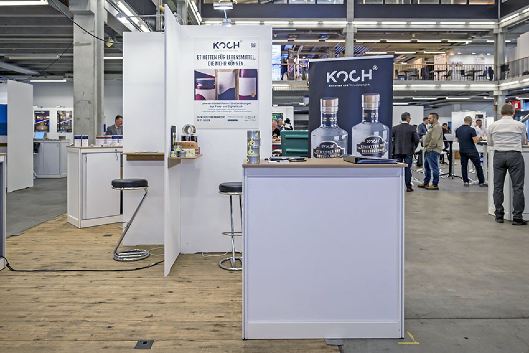 Messestand für Koch mit Beschilderung, auf der Etiketten für Lebensmittelprodukte und Muster von Glasflaschen auf einer Theke ausgestellt sind.